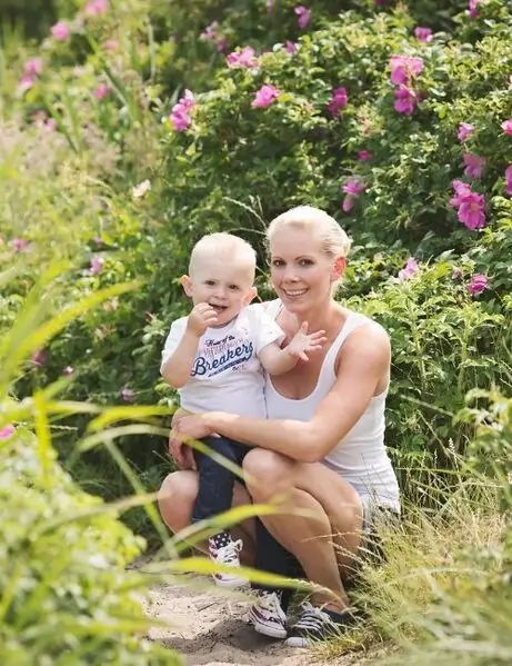 Elke Lengauer crouching outdoors in a grassy area with flowers, holding a small child who is also smiling.