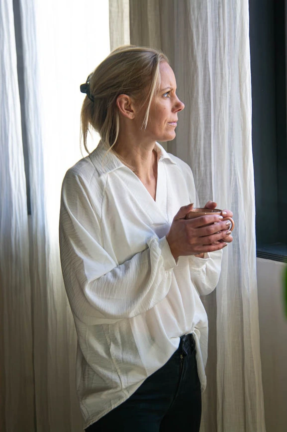 Elke Lengauer in a white long-sleeved shirt standing by a window, holding a mug and looking to the side.